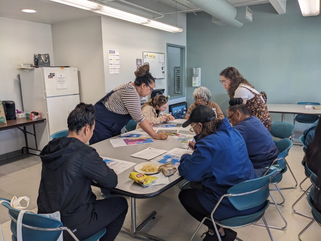 A group of people gather around a table indoors with maps as part of a shoreline planning workshop
