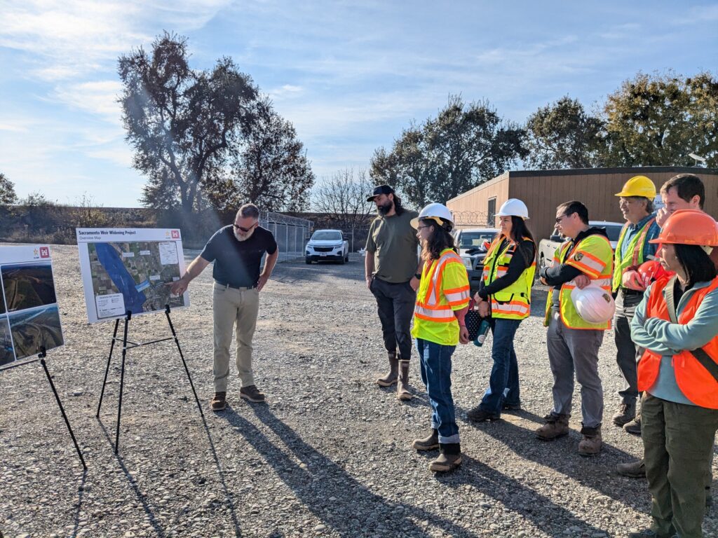 A group of people in hard hats and high-visability vests gather To hear a presentation about the Sacramento Weir widening project
