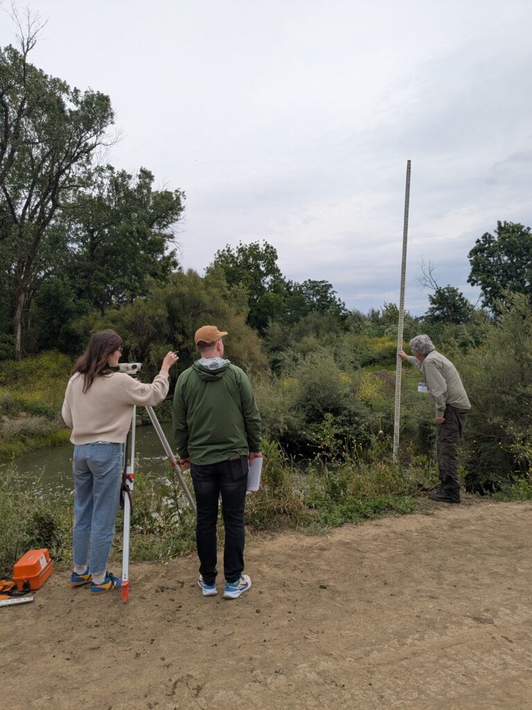 Two people stand in front of a surveying auto level and point at a 3rd person holding a stadia rod while performing a fish passage assessm while performing a fish passage assessment