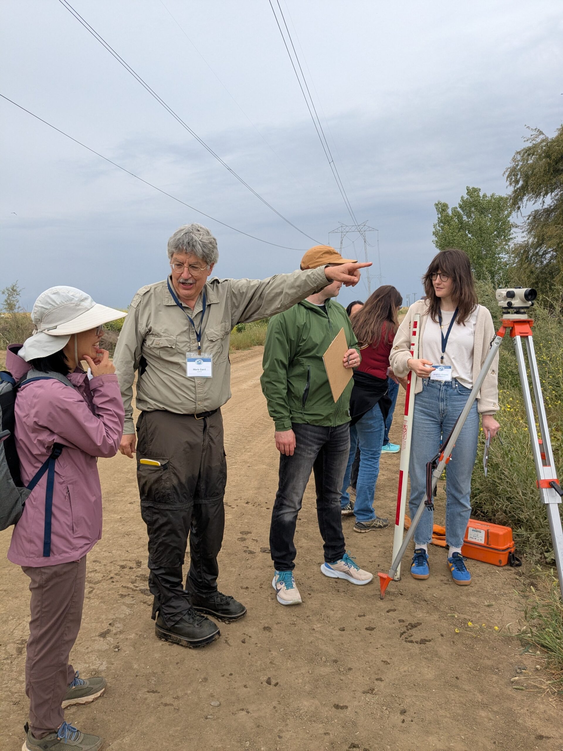 Two people stand in front of a surveying auto level and point at a 3rd person holding a stadia rod while performing a fish passage assessm while performing a fish passage assessment
