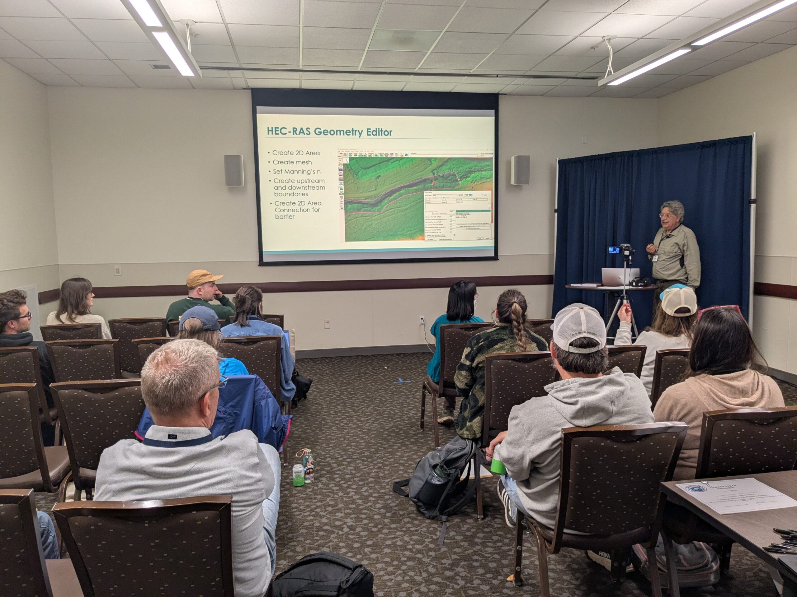 A group of people observes a presentation in a meeting, learning how to model hydrography in a modeling software.