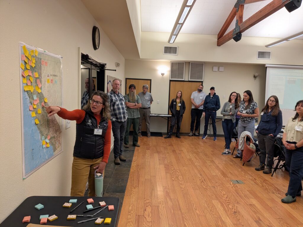 A person stands in front of a map of California covered with colored sticky notes and points to a as a large group watches
