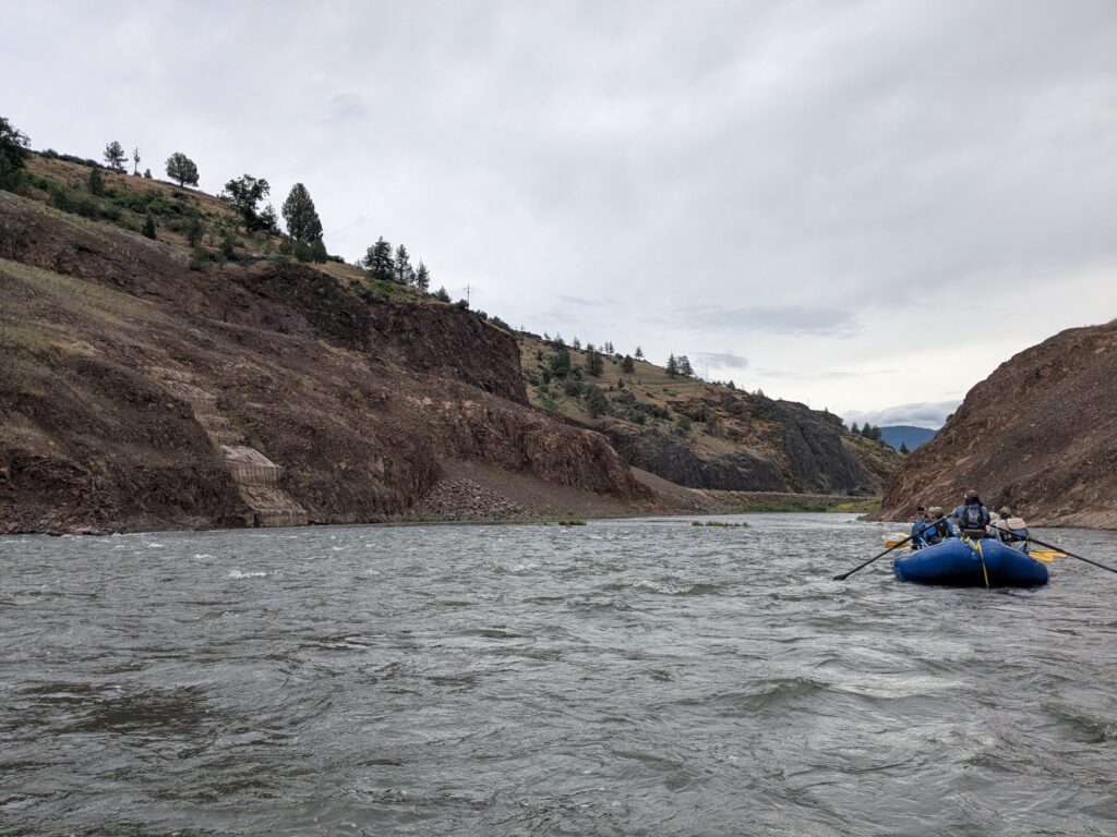 A raft flows on the Corlamoth River with a few of the core of the Iron Gate Dam embedded in the hillside